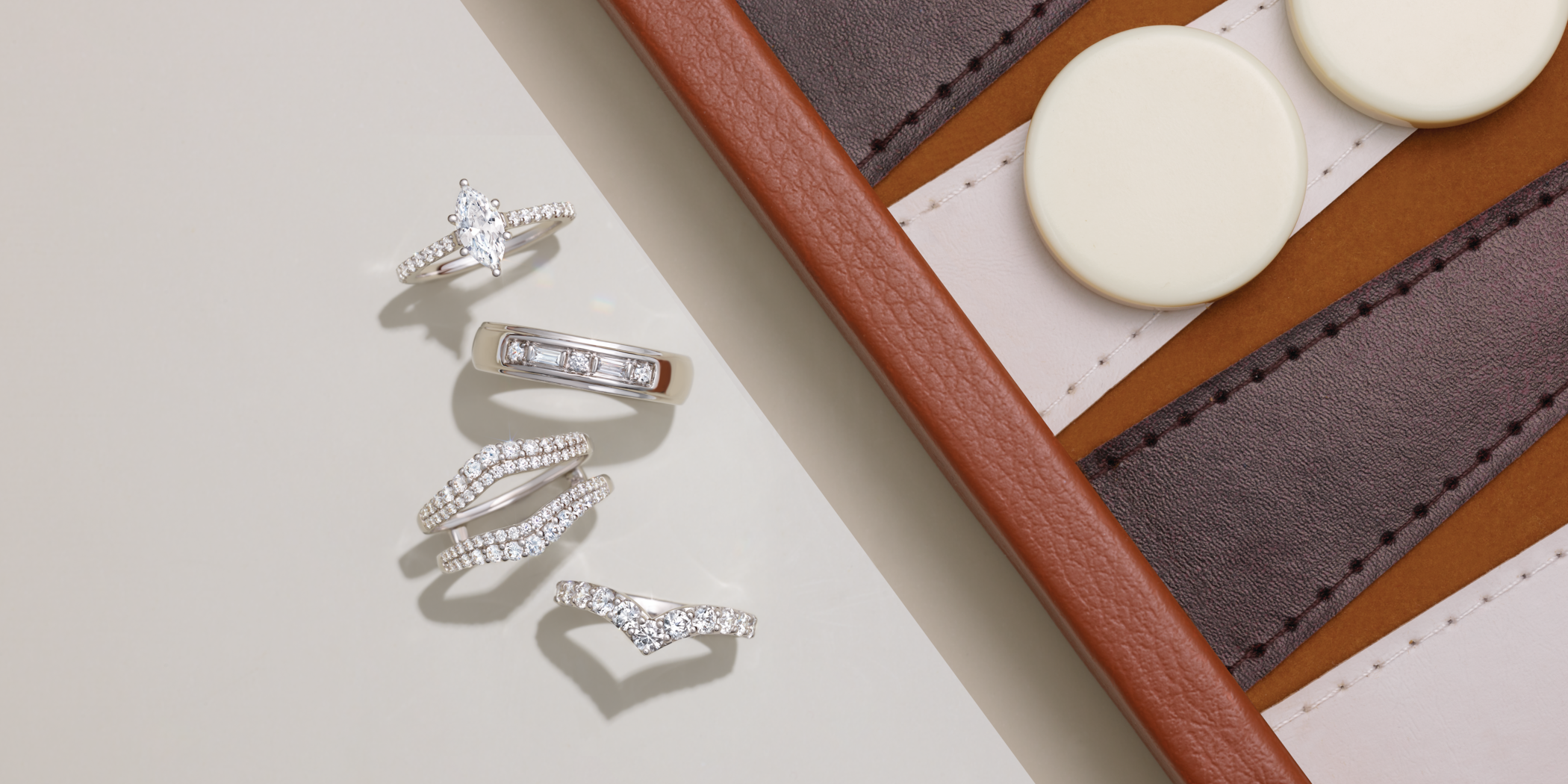 Five diamond rings, including a marquise engagement ring and baguette band, arranged by a leather backgammon board. Custom jewelry by Bentzion in Airmont, NY.