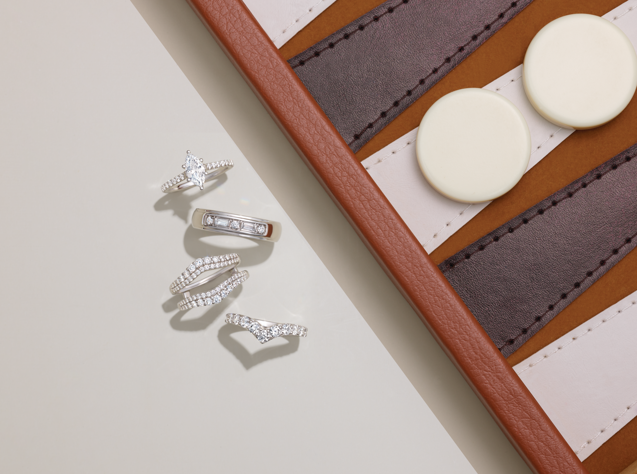 Five diamond rings, including a marquise engagement ring and baguette band, arranged by a leather backgammon board. Custom jewelry by Bentzion in Airmont, NY.