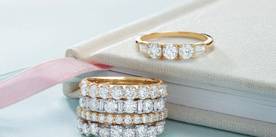 A stack of four diamond eternity bands in gold and platinum alongside a yellow gold three-stone diamond ring resting on an off-white fabric journal at Motek Jewelry Design Studio in Airmont, NY.