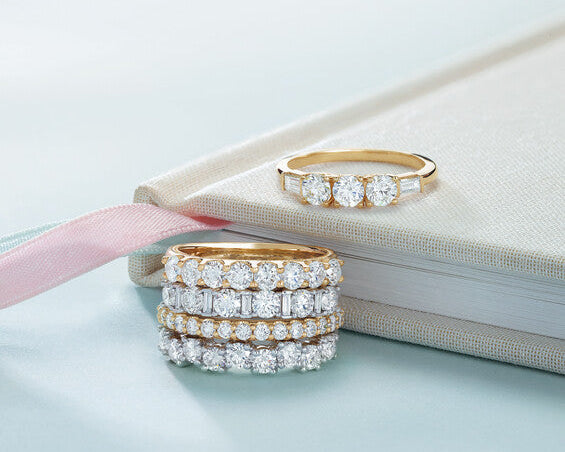 A stack of four diamond eternity bands in gold and platinum alongside a yellow gold three-stone diamond ring resting on an off-white fabric journal at Motek Jewelry Design Studio in Airmont, NY.