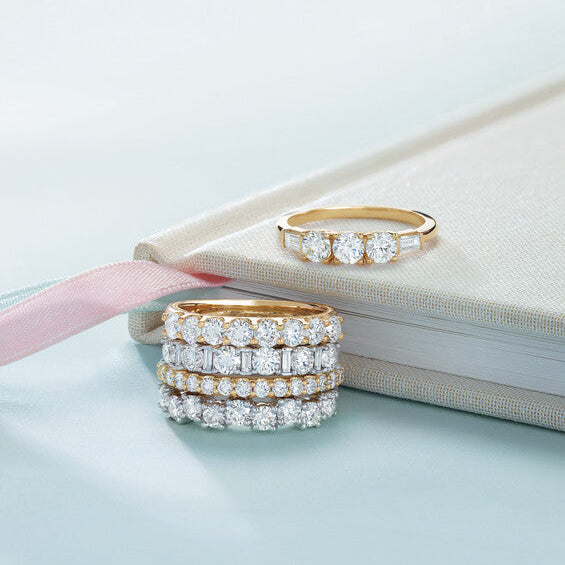 A stack of four diamond eternity bands in gold and platinum alongside a yellow gold three-stone diamond ring resting on an off-white fabric journal at Motek Jewelry Design Studio in Airmont, NY.