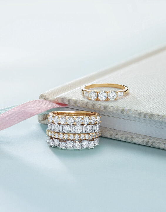 A stack of four diamond eternity bands in gold and platinum alongside a yellow gold three-stone diamond ring resting on an off-white fabric journal at Motek Jewelry Design Studio in Airmont, NY.
