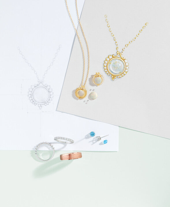 Custom jewelry design sketches, technical CAD blueprints, opal pendant necklaces, diamond eternity bands, and loose gemstones on a design desk at Motek Jewelry in Airmont NY