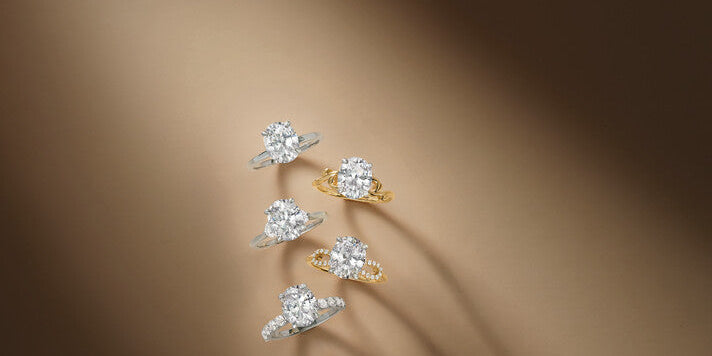 Five oval diamond solitaire engagement rings arranged vertically on a neutral brown backdrop, featuring various platinum and 18k yellow gold bands with pavé details, at Motek Jewelry Design Studio in Airmont, NY