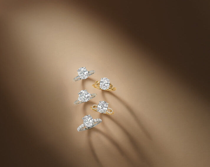 Five oval diamond solitaire engagement rings arranged vertically on a neutral brown backdrop, featuring various platinum and 18k yellow gold bands with pavé details, at Motek Jewelry Design Studio in Airmont, NY