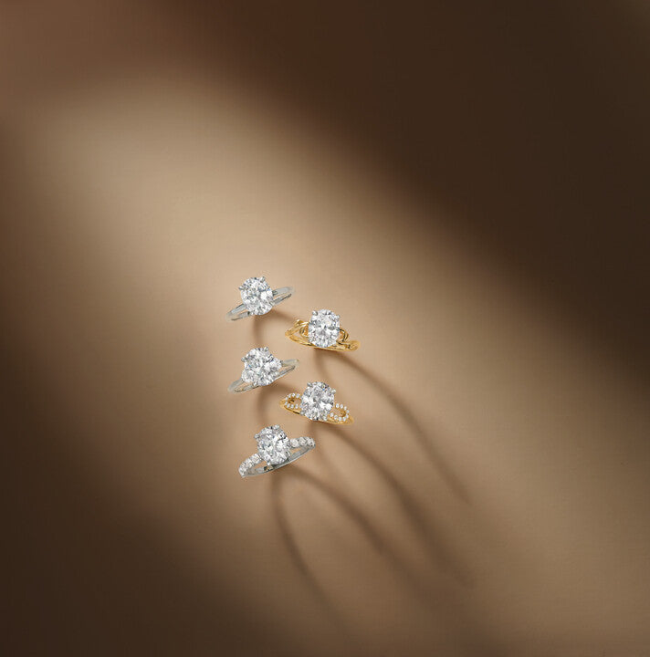 Five oval diamond solitaire engagement rings arranged vertically on a neutral brown backdrop, featuring various platinum and 18k yellow gold bands with pavé details, at Motek Jewelry Design Studio in Airmont, NY