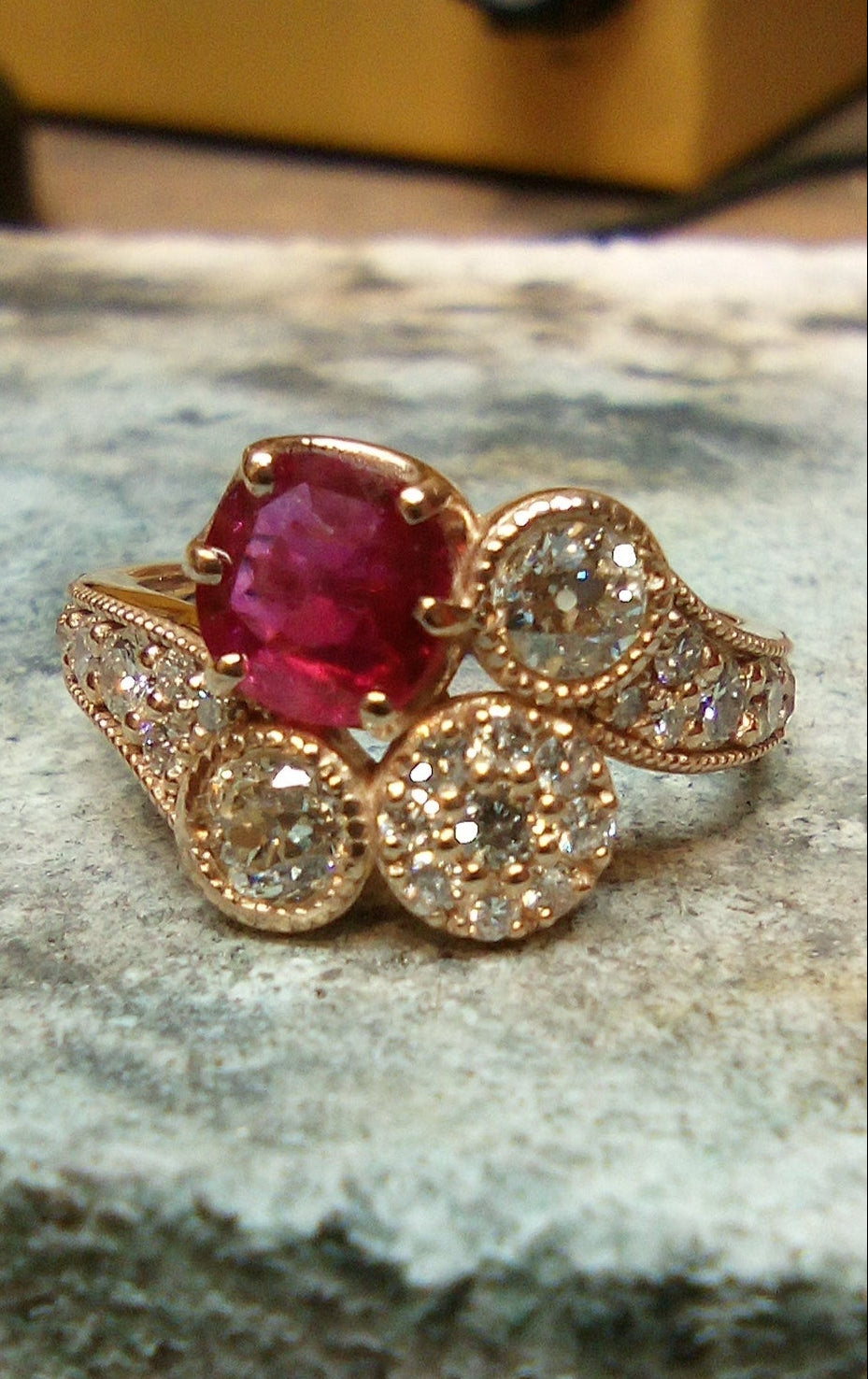 Yellow gold ring with a red gemstone on a textured surface, a soldering block on a jewelers bench. 